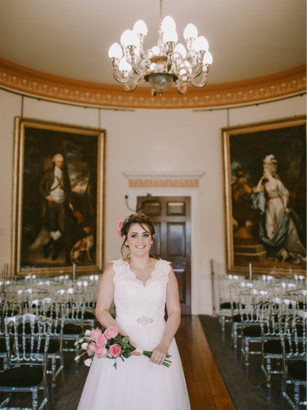Bride holding bouquet of pink roses stood in the dining room, with ceremony seating layout.