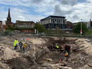 A view of the archaeological dig at Forge Island. Residents look on as archaeologists work in a trench. Historical walls and foundations can be seen.