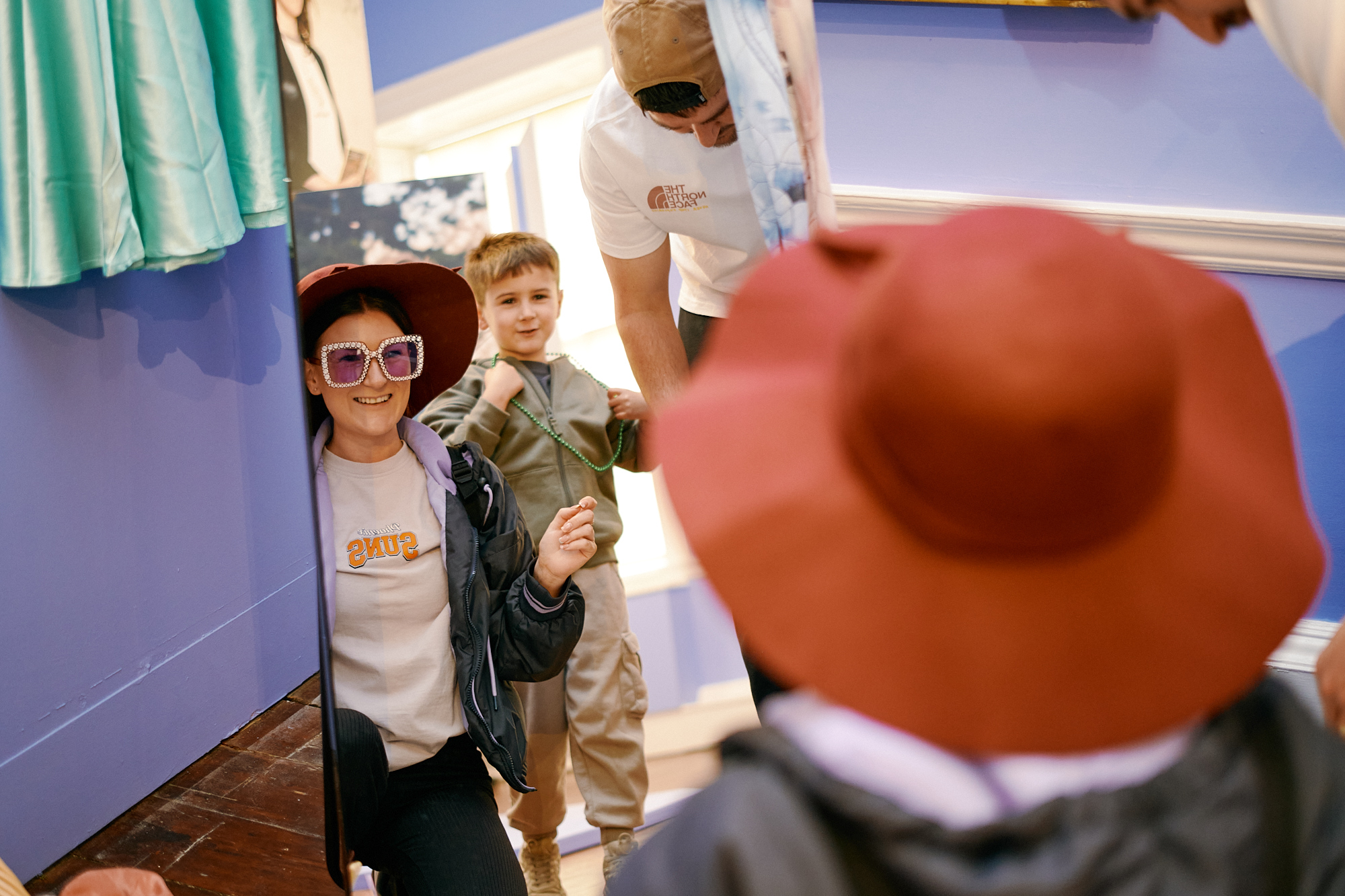 A woman in a big hat and glasses knelt down in front of a mirror. A child and a man are  behind her.