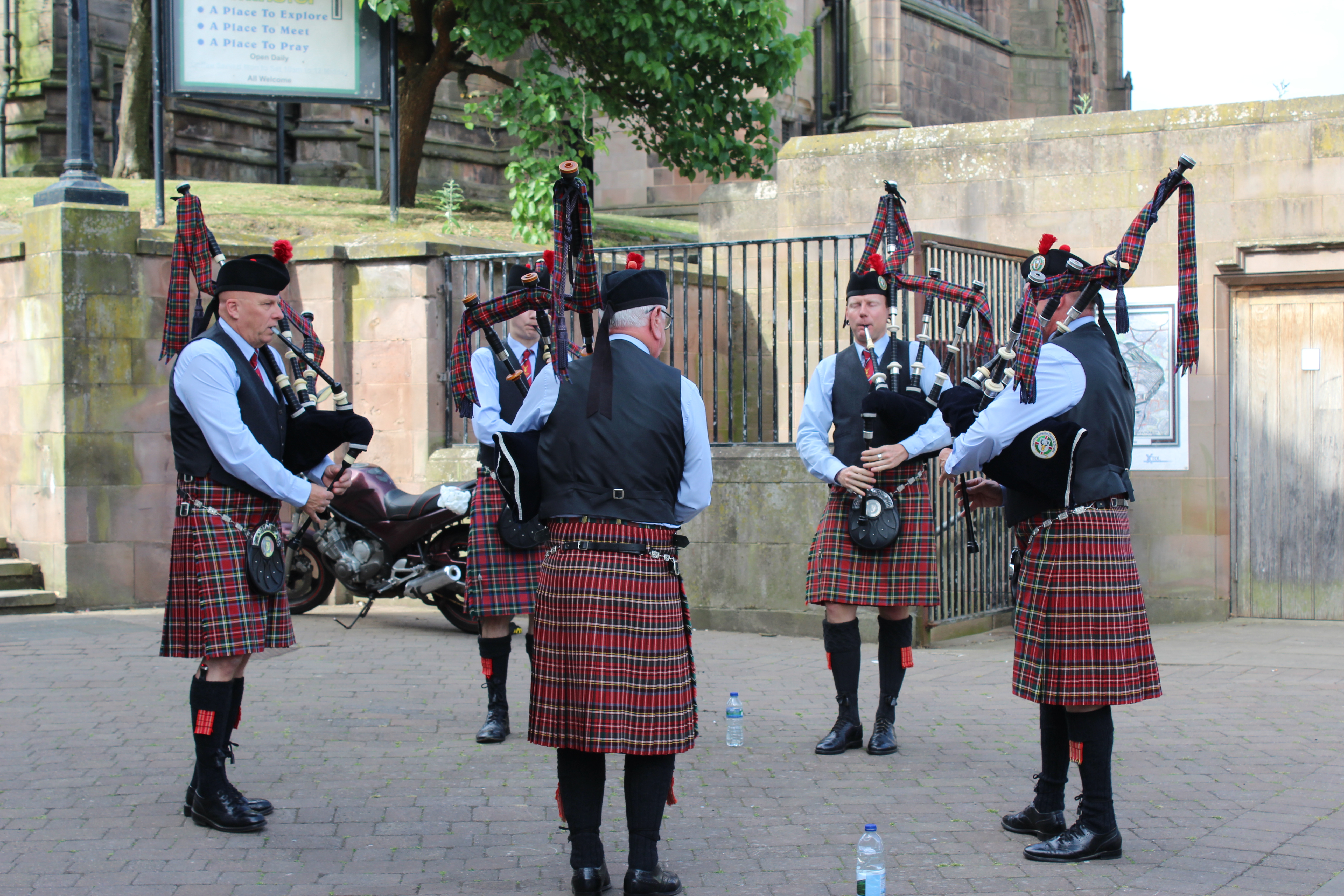 Five bagpipers in traditional Scottish attire, including kilts and black hats, perform in a circle on a paved area. 