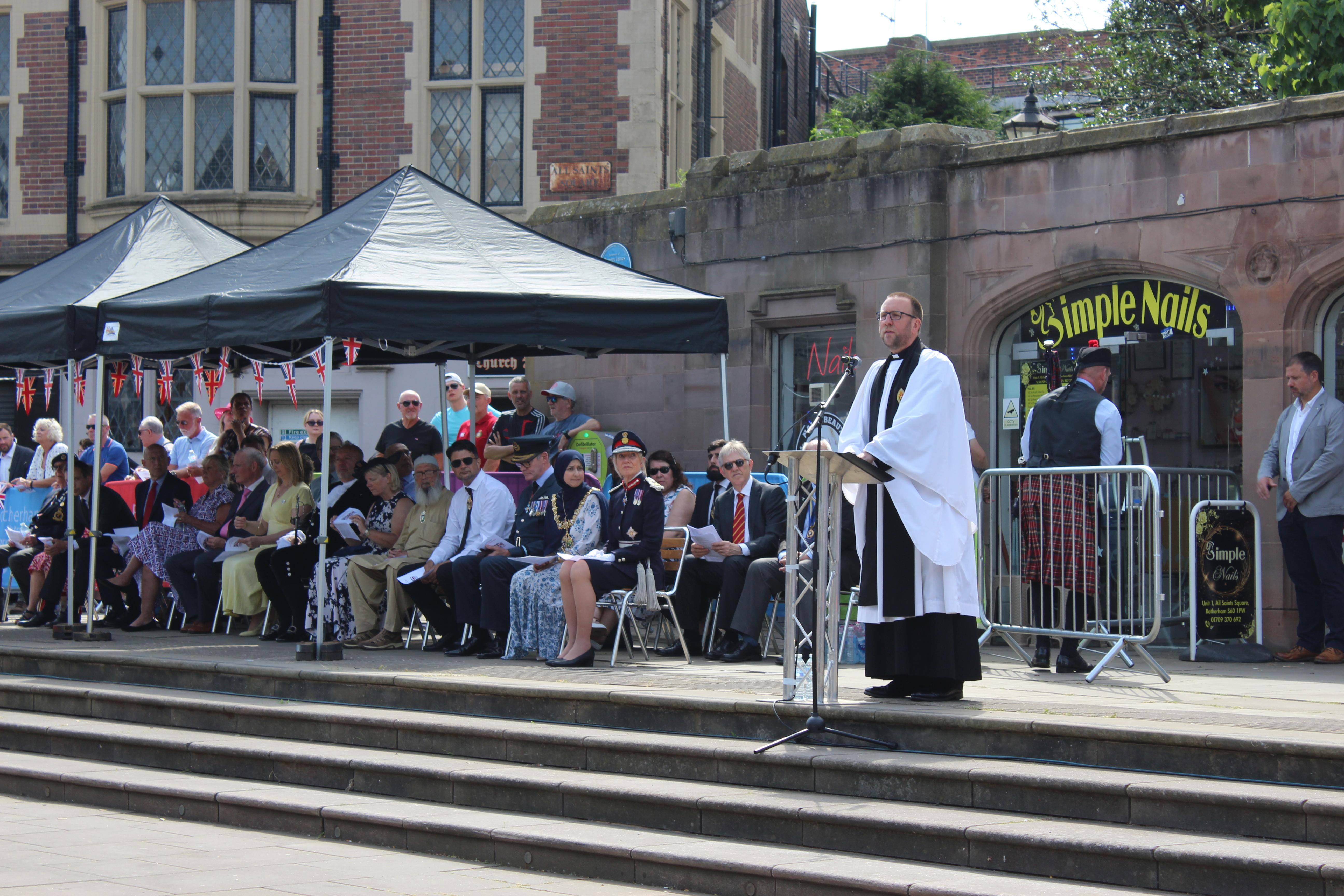 An outdoor public event in front of a historic building, with attendees seated under black canopies. A person in religious attire speaks at a podium on the steps.