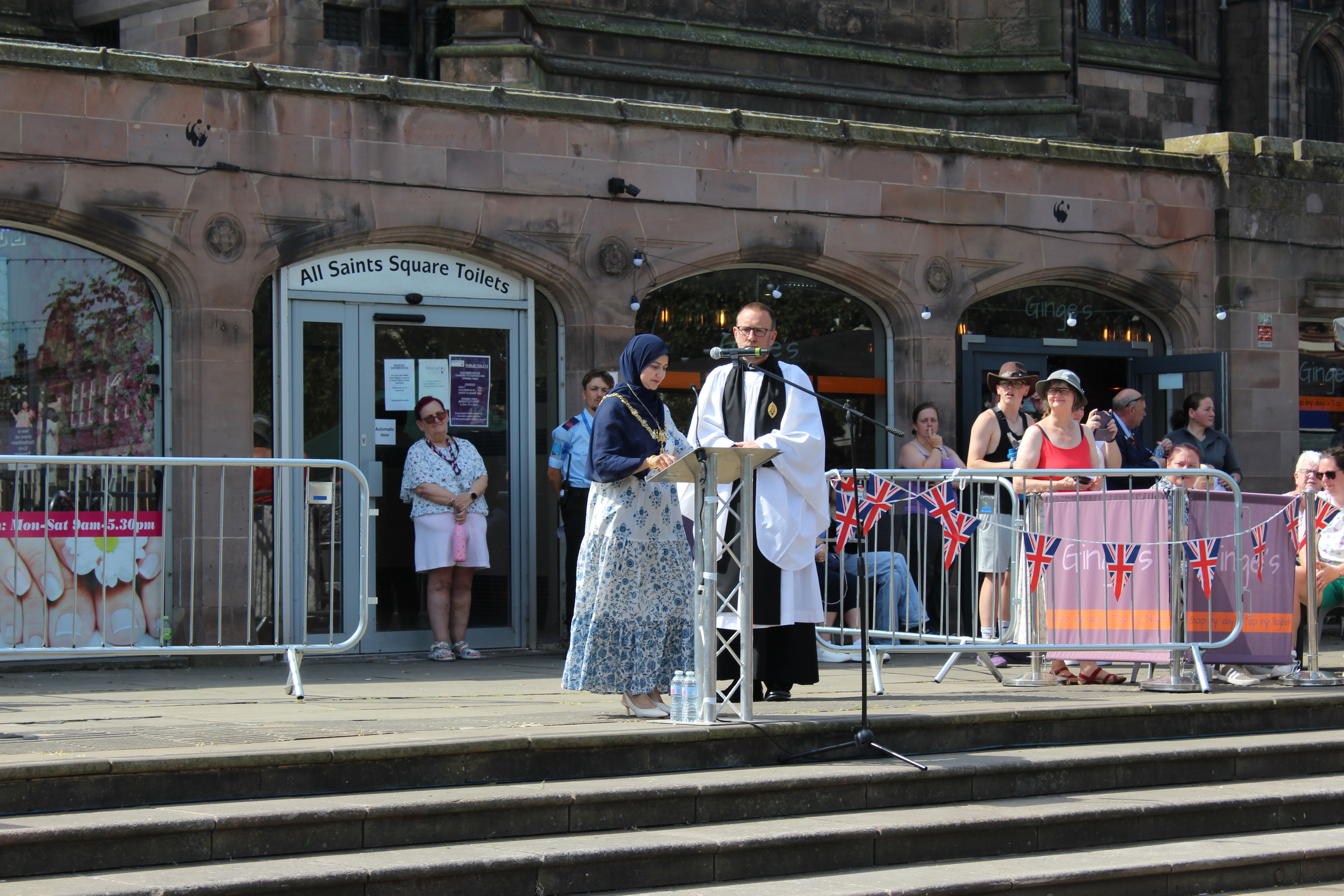 Two speakers at a podium during an outdoor event in All Saints Square. Attendees are gathered behind Union Jack-decorated barriers.