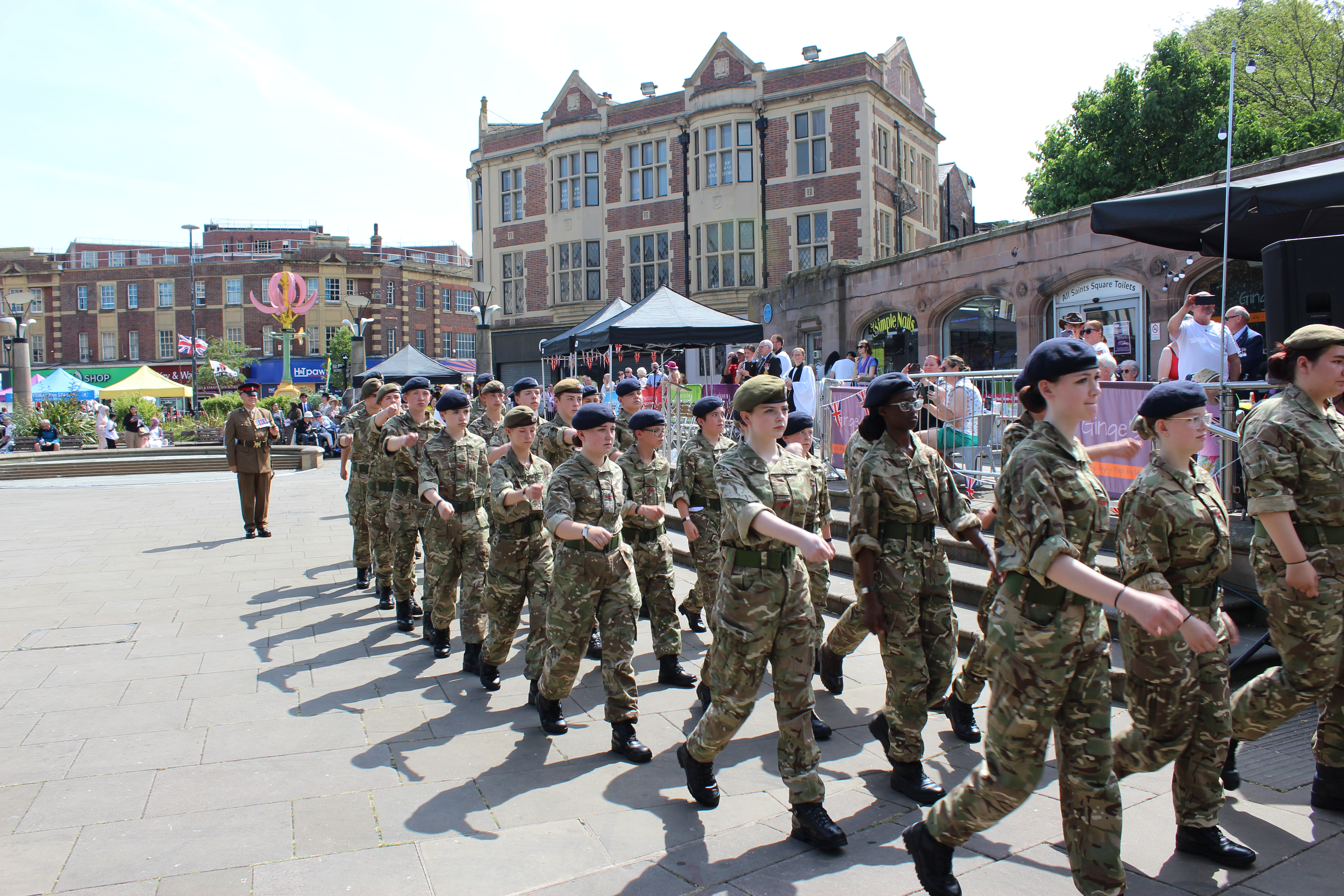 A group of people in military uniforms marches in formation on a paved area. Spectators watch from behind barriers, with tents and stalls visible in the background on a sunny day.