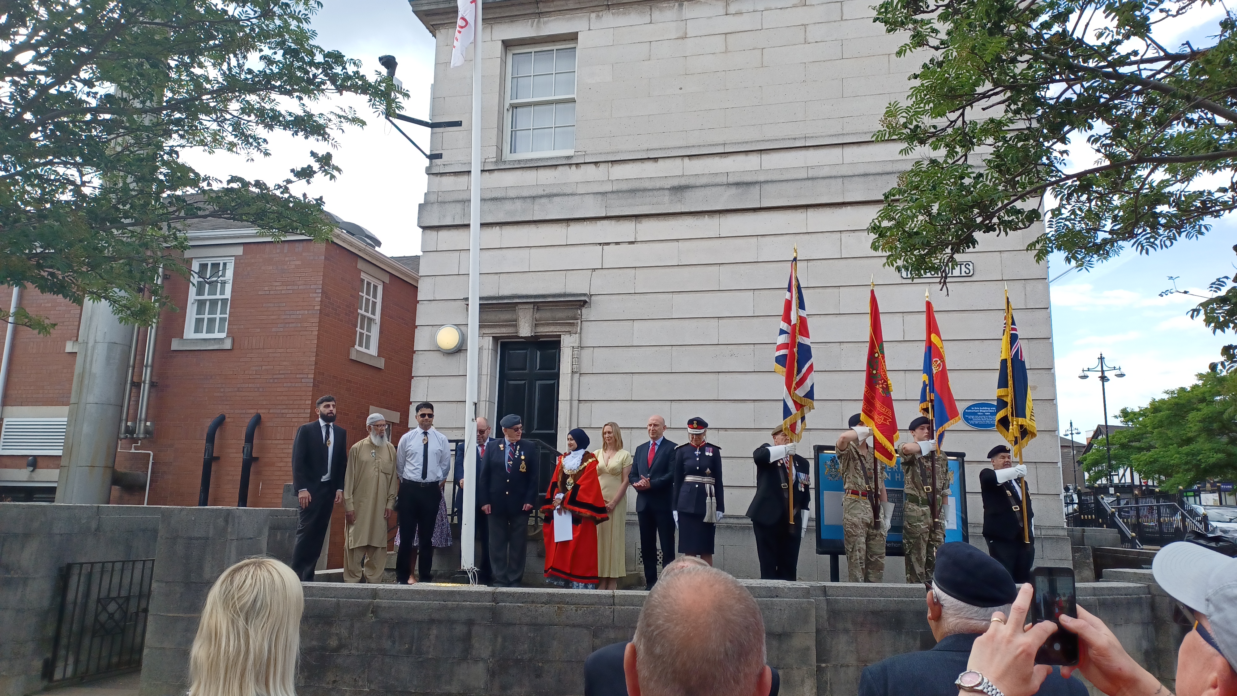 Group of individuals in military uniforms, traditional robes, and formal attire standing on a raised platform in front of a historic building with a flagpole. Several people hold flags, while an audience watches and takes photos.