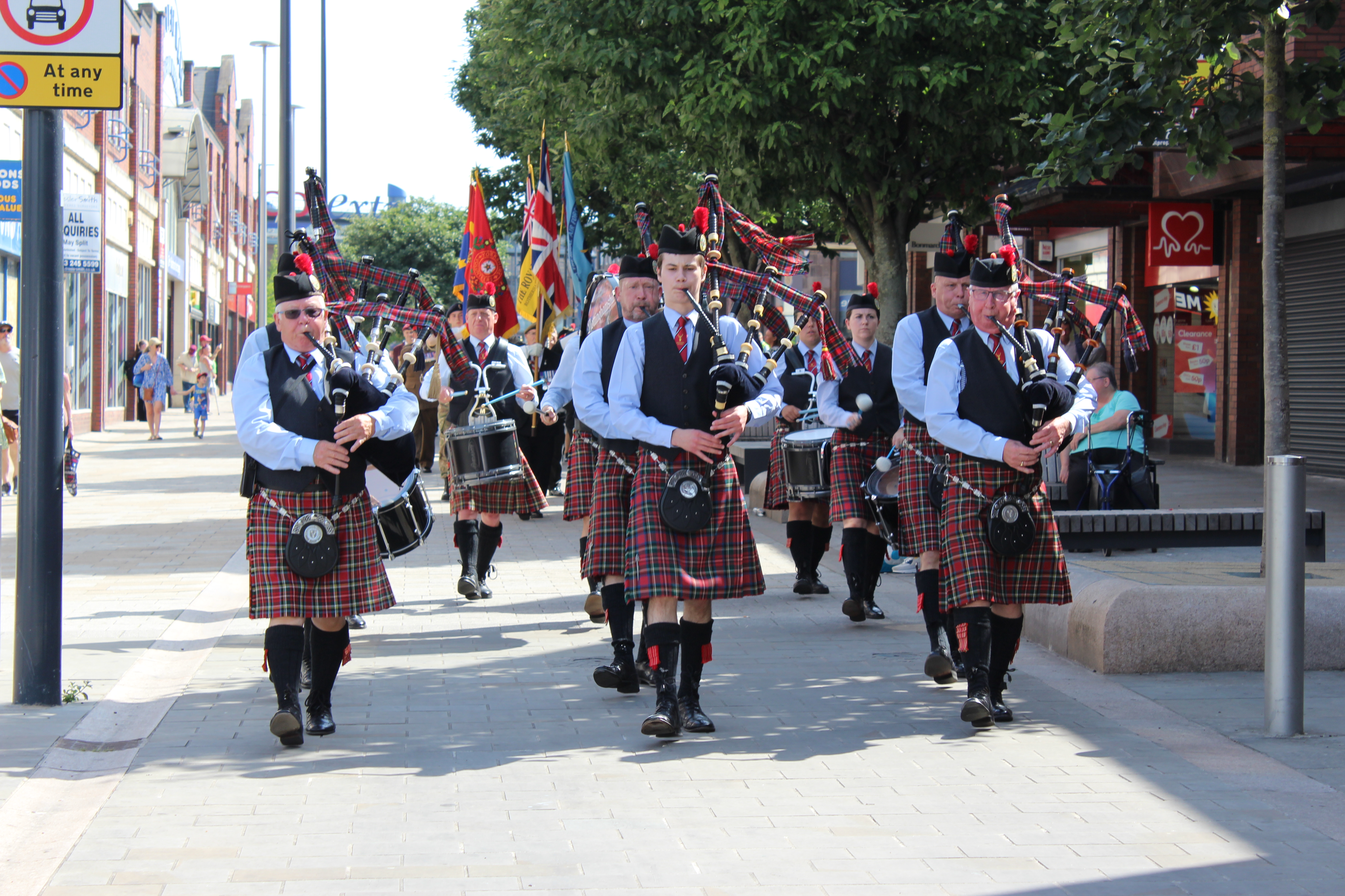 Group in traditional Scottish attire, including kilts and sporrans, marching down a street while playing bagpipes and drums. 