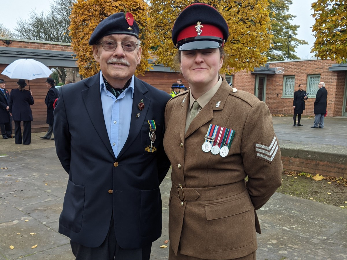 Female soldier in brown dress uniform wearing medals, with older man wearing medals on his navy blue suit jacket and a poppy on his black beret