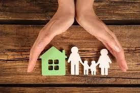 Hands protecting paper cut-outs of a family and a green house on a wooden table