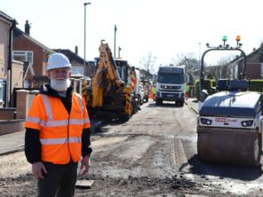 Cllr Taylor stood on a road in a hard hat and hivis while road works are ongoing behind.