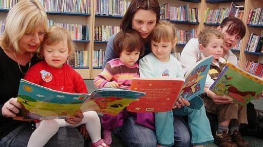 Three people in a library with a toddler on their knee, reading a book.