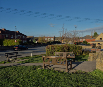 Two benches opposite each other with a stone path in the middle. Grass sections behind each one with houses in the background.