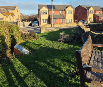 A grass section with a bench to the left with a hedge on the right and houses in the background.