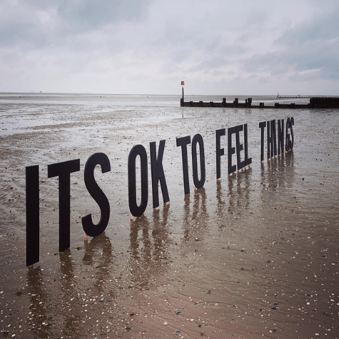 A beach with the phrase 'ITS OK TO FEEL THINGS' placed on it in large letters.