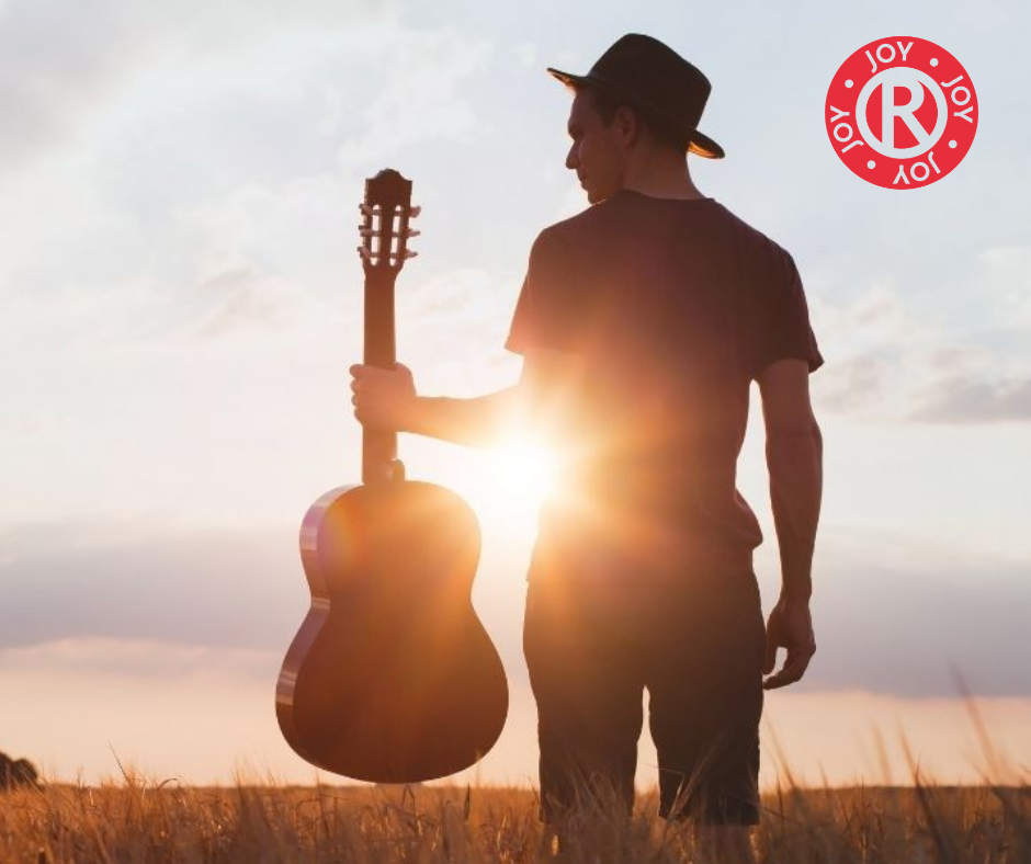 A man holding a guitar in front of a sunset. The picture has the word Joy written on it.