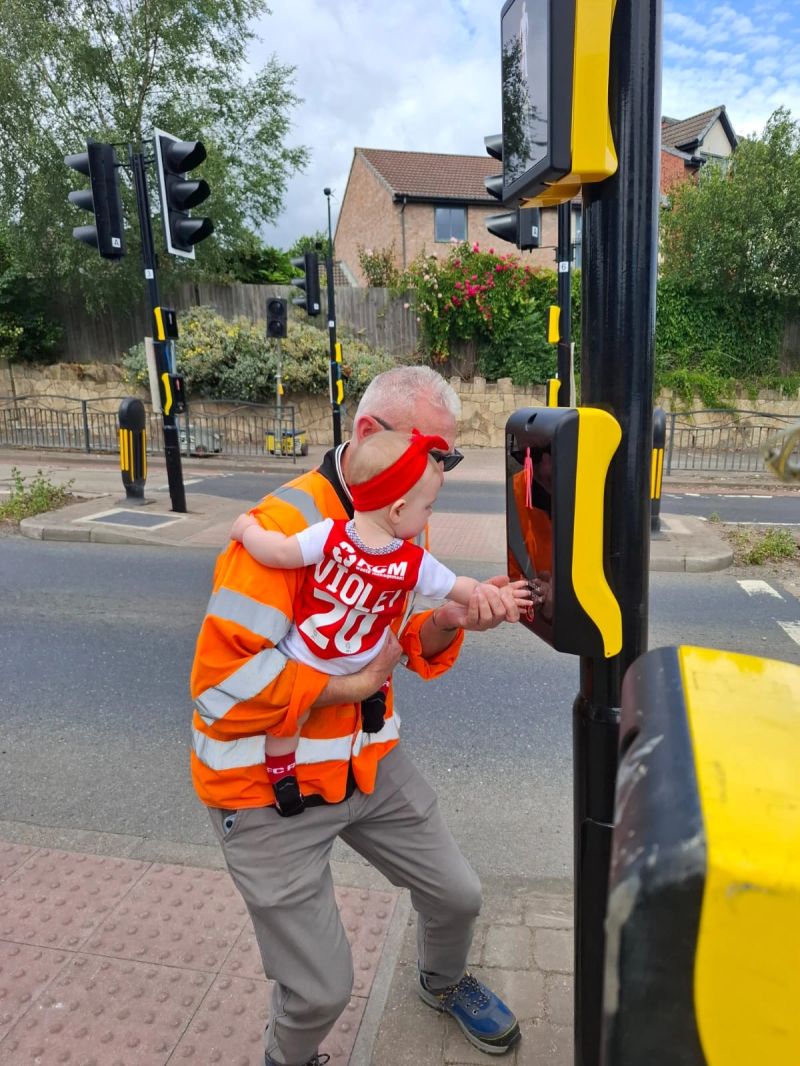 A man in an orange high-visibility jacket helps a baby press a button at a pedestrian crossing for the Dale Road junction.